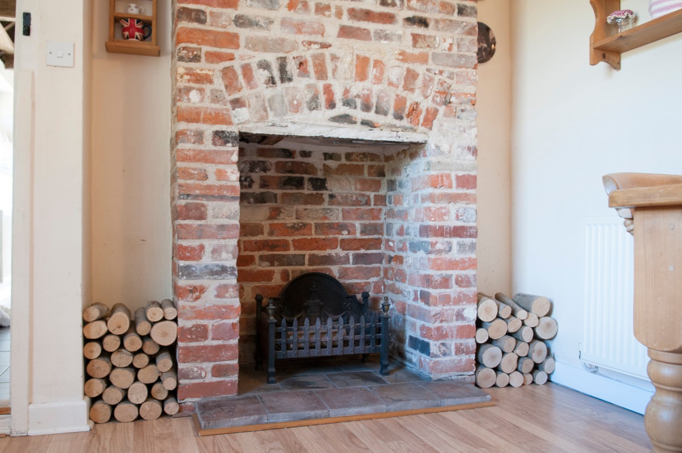 A view of a wrought iron grate in a bare brick, rustic fireplace with chopped logs stacked up to the sides.