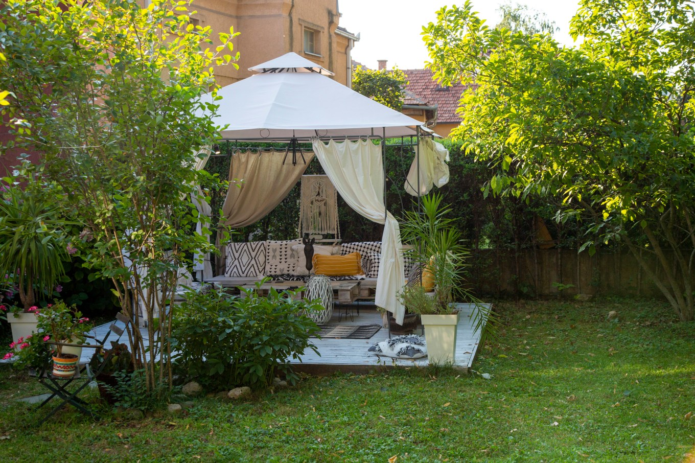 Beautiful cozy gazebo with fabric curtains in the backyard surrounded with trees, flowers, lawn and fence