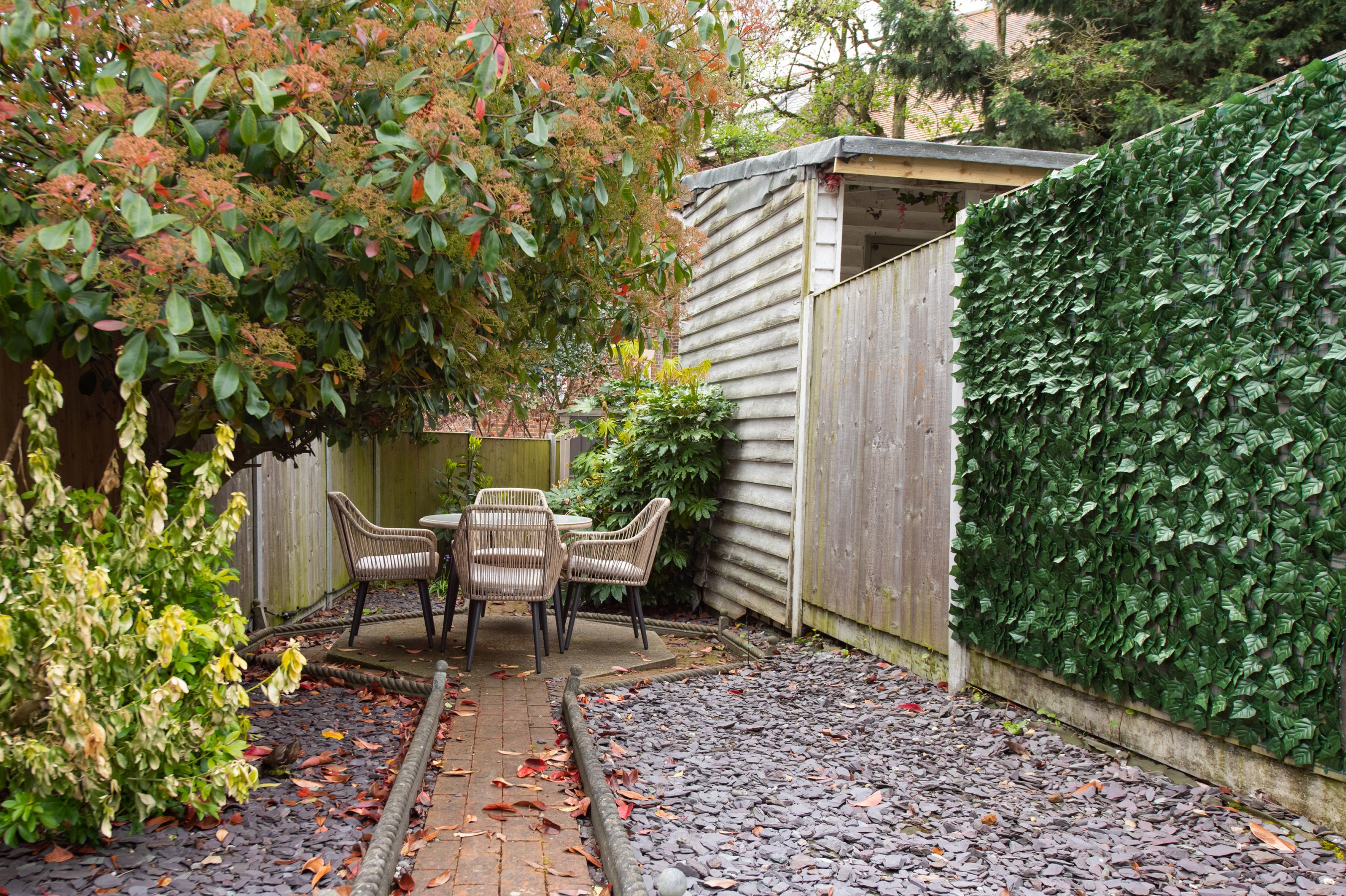 A general view of a small courtyard garden hidden from view by a greenery panel with a cobblestone patio, slate chippings, table and string rope chairs