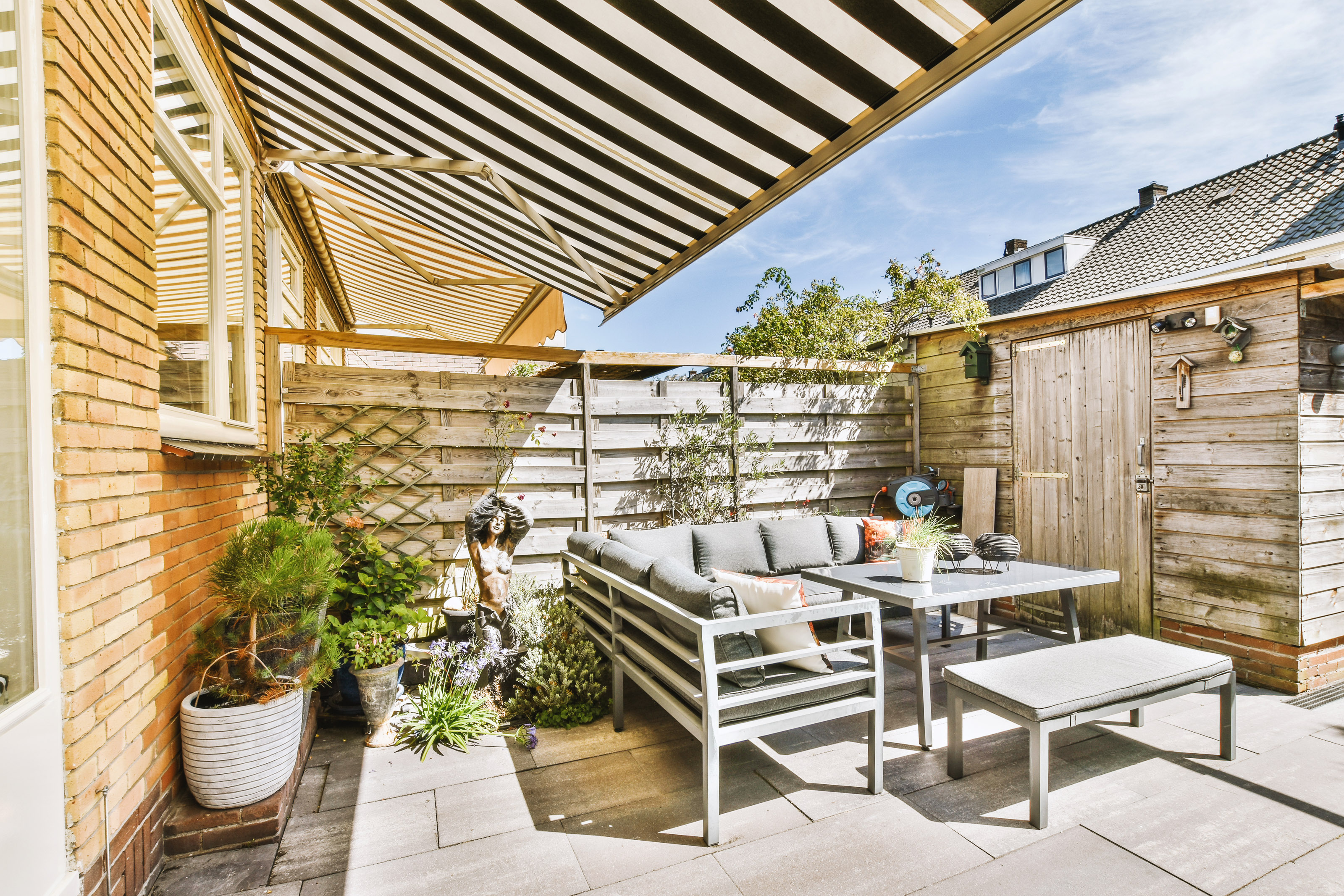 Neat paved patio with sitting area covered by a retractable awning and small garden near wooden privacy fence
