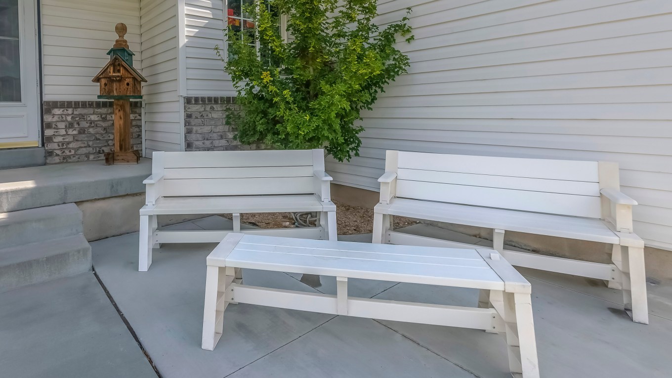 Home with white wooden furniture on front patio. Home with wooden benches and table on the patio connected to stairs leading to the front door. A small tree against the wall is behind the furniture.