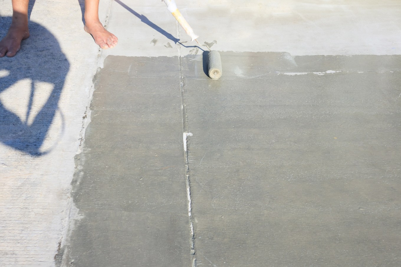 Someone using a roller painter to seal and paint a concrete pavement floor.