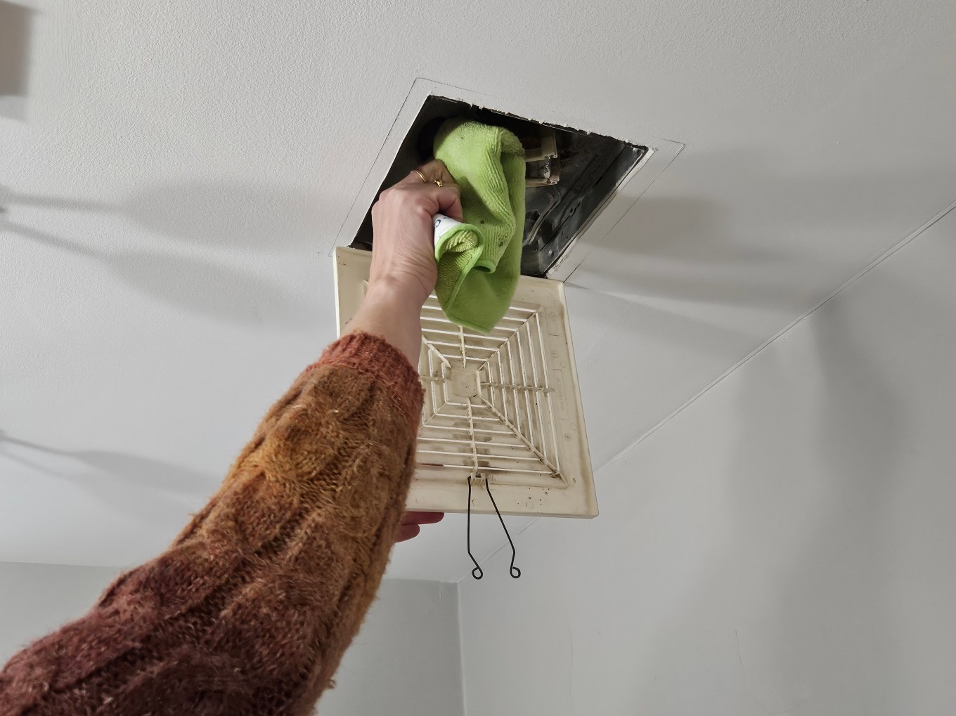 A person cleaning the vent fan in a bathroom with a microfiber cloth.