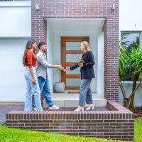 Real estate agent showing a couple a new house. The contemporary house is an off-white and has a brick facade.