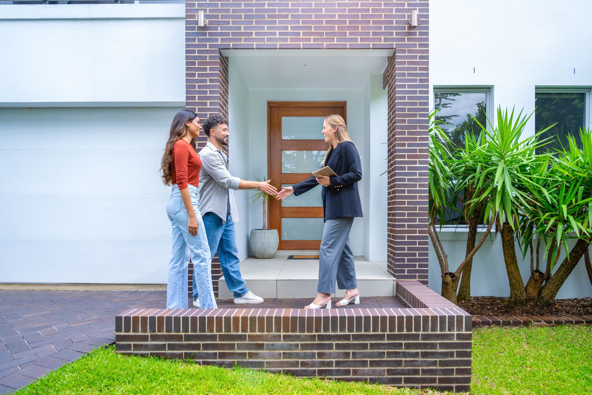 Real estate agent showing a couple a new house. The contemporary house is an off-white and has a brick facade.