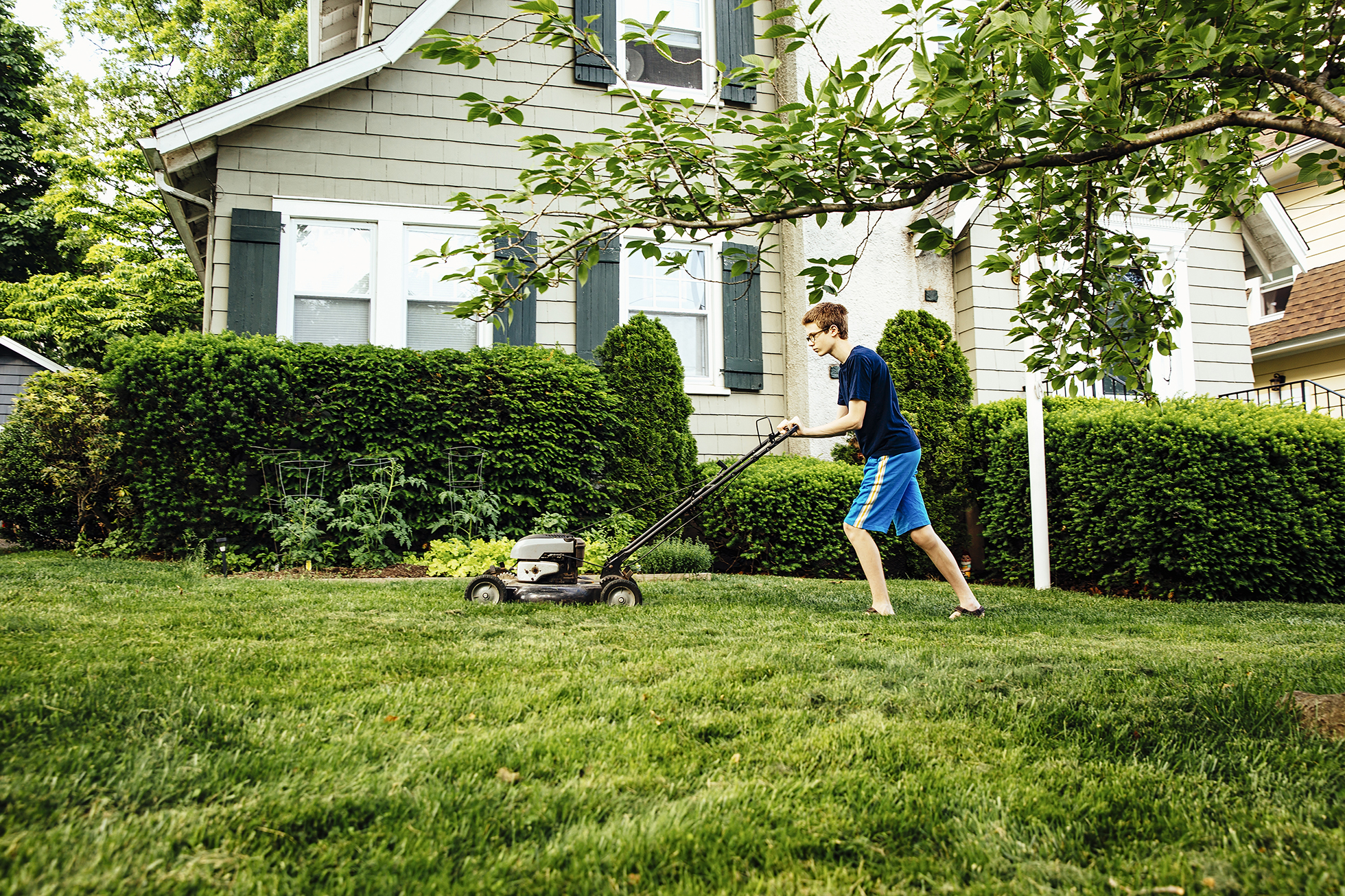 Tween boy in blue mowing lawn outside light green house