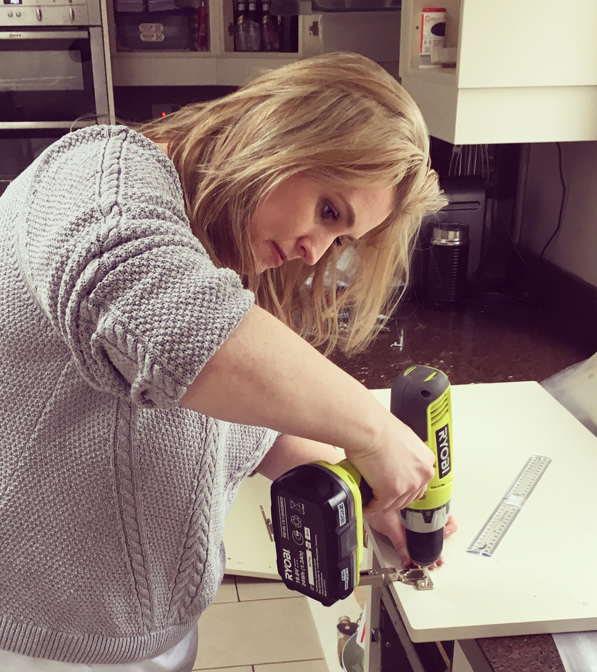 Blond woman in gray sweater assembling cabinet with drill