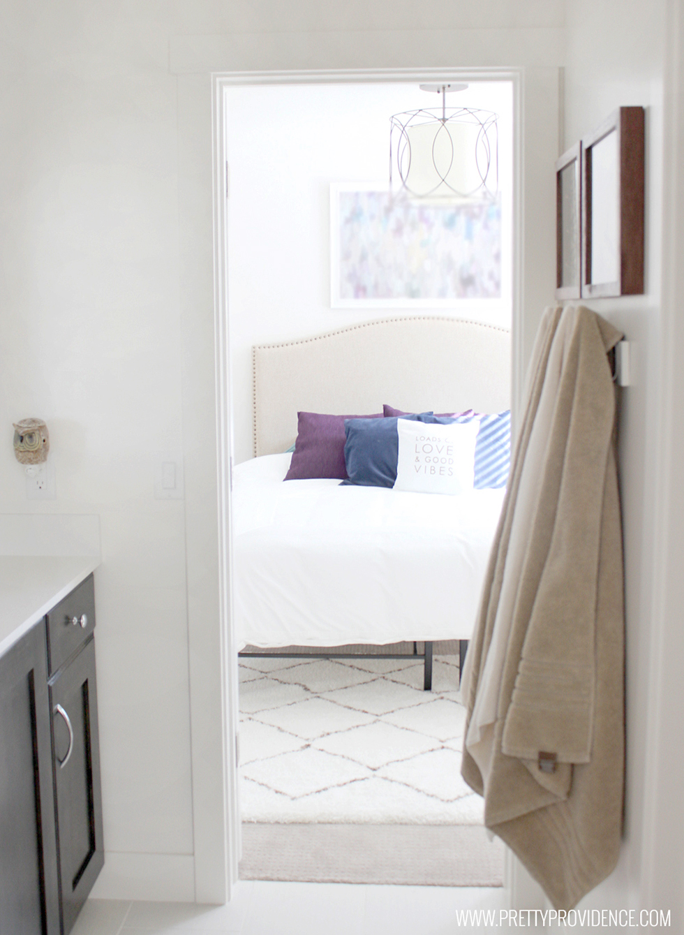 Glimpse from bathroom of white bed with blue & purple pillow