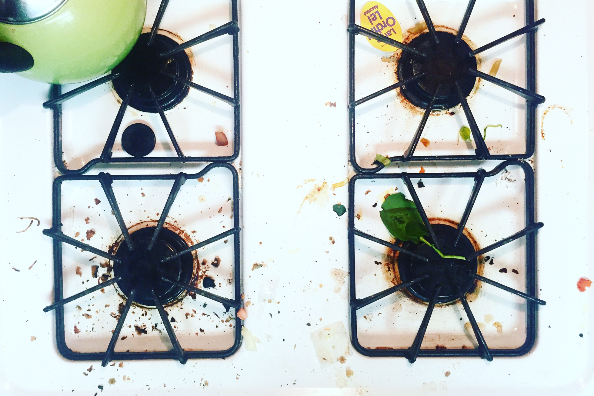 A gas stovetop with food crumbs, green teapot
